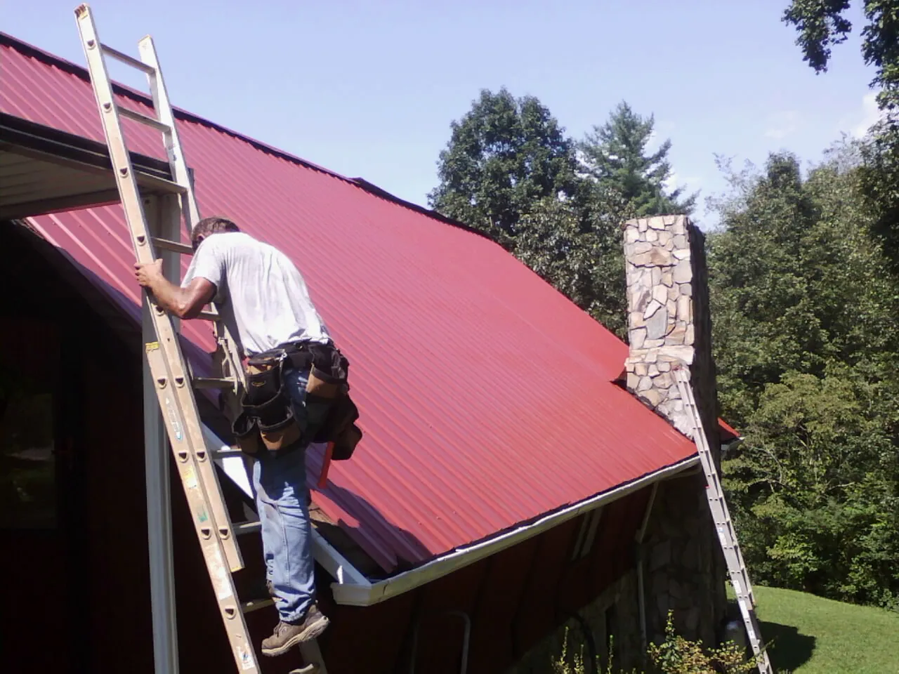 Roofer on a ladder beside a red metal roof and stone chimney — Soffit & Fascia Repair by Roofing Brothers in Altaville